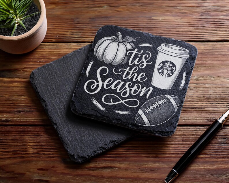 May include: Two square black slate coasters. One coaster has a pumpkin, coffee cup, and football design with the text "'tis the Season" in white. The other coaster is plain. A pen rests on the wooden surface.