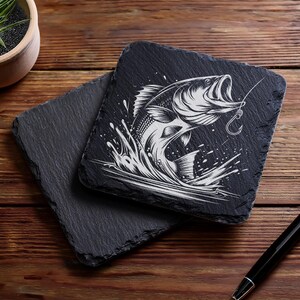 May include: Two square black slate coasters. One coaster features a detailed white illustration of a fish jumping out of water with a fishing hook. The other coaster is plain. A pen and a small potted plant are also visible.