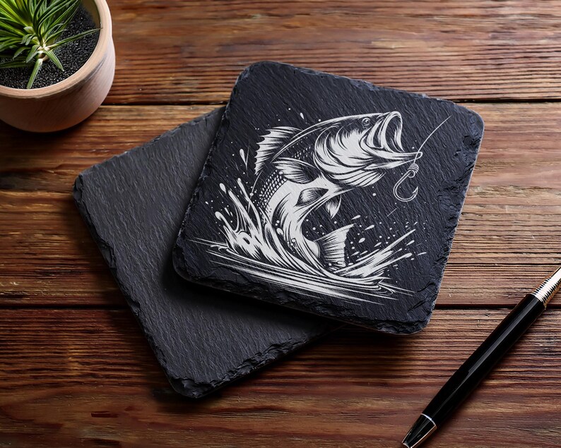 May include: Two square black slate coasters. One coaster features a detailed white illustration of a fish jumping out of water with a fishing hook. The other coaster is plain. A pen and a small potted plant are also visible.