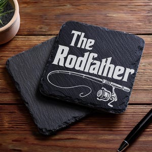 May include: Two square, dark gray slate coasters. One coaster has the text "The Rodfather" with a fishing rod graphic. A small potted plant and a pen are also in the image.