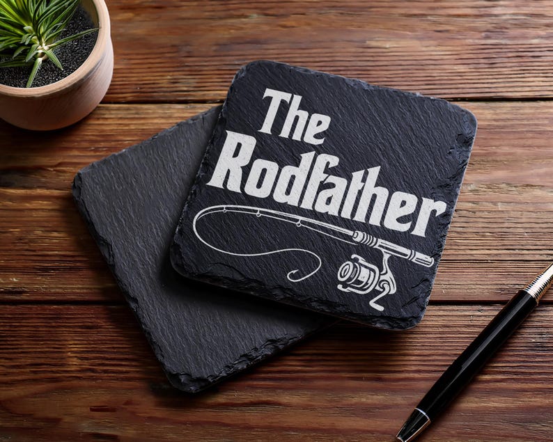 May include: Two square, dark gray slate coasters. One coaster has the text "The Rodfather" with a fishing rod graphic. A small potted plant and a pen are also in the image.
