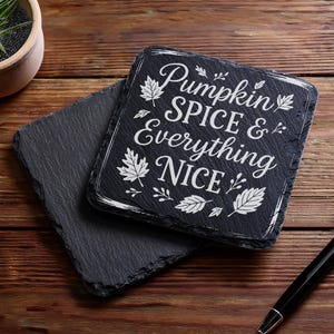 May include: Two dark gray slate coasters on a wooden surface. One coaster has the phrase "Pumpkin Spice & Everything Nice" in white script and leaf designs. A black pen rests nearby, and a small potted plant is in the background.
