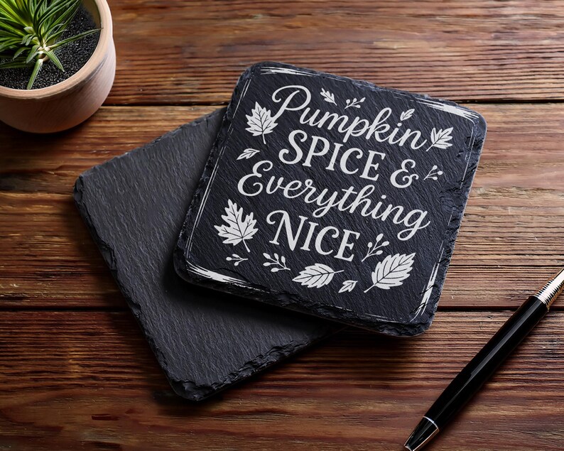 May include: Two dark gray slate coasters on a wooden surface. One coaster has the phrase "Pumpkin Spice & Everything Nice" in white script and leaf designs. A black pen rests nearby, and a small potted plant is in the background.