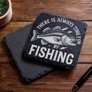 May include: Two square black slate coasters. One coaster has the text "THERE IS ALWAYS TIME FOR FISHING" with a fish illustration. The other coaster is plain. A black pen rests on the wooden surface.
