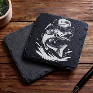 May include: Two square black slate coasters. One coaster features a white graphic of a fish wearing sunglasses and a hat that says "FISHING." The other coaster is plain. The coasters are on a wooden surface.