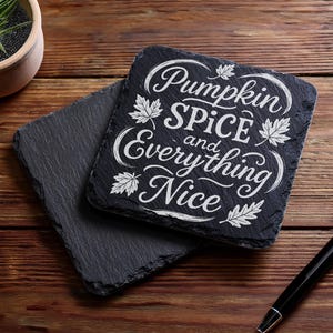 May include: Two black slate coasters with the text "Pumpkin Spice and Everything Nice" in white script. The coasters are square with rough edges and sit on a wooden surface. A pen and a small potted plant are also visible.