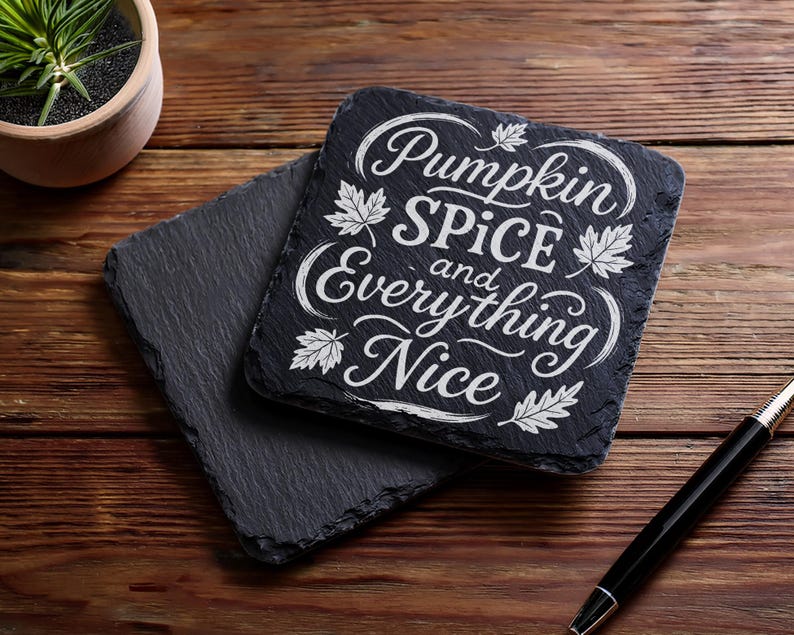 May include: Two black slate coasters with the text "Pumpkin Spice and Everything Nice" in white script. The coasters are square with rough edges and sit on a wooden surface. A pen and a small potted plant are also visible.
