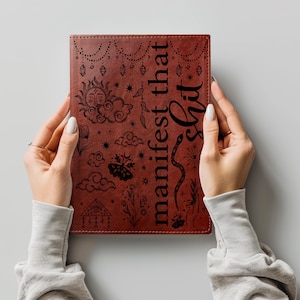 May include: A rectangular brown notebook featuring black line art illustrations of a sun, clouds, a moth, a snake, and crystals. The text "manifest that shit" is printed vertically in a bold, black serif font on the right side of the cover.