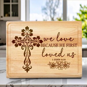 May include: A rectangular wooden cutting board with a decorative cross design and the text "we love because he first loved us" and "1 John 4:19". The cross is adorned with floral accents.