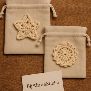 Puede incluir: Dos bolsas beige con cordón y adornos de ganchillo. Una bolsa presenta una estrella, la otra un diseño circular. Una tarjeta de visita con el texto "BijAlunaStudio" descansa debajo de las bolsas sobre una superficie de madera.