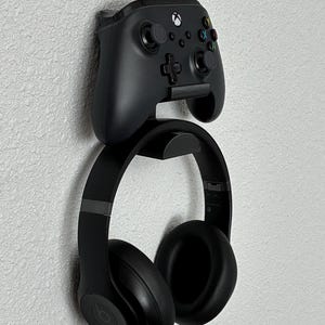 May include: A black wall-mounted gaming accessory holder. The holder features a top section designed to hold a black Xbox controller and a lower section for black over-ear headphones. The holder is mounted on a textured white wall.