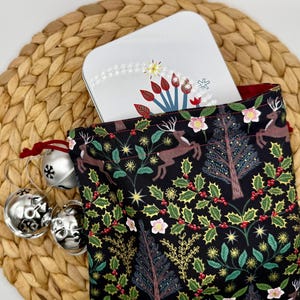 Puede incluir: Una bolsa negra con cordón con un patrón festivo de ciervos, acebo y árboles, que contiene una lata rectangular blanca con un diseño navideño. Campanas plateadas con cintas rojas están al lado de la bolsa, sobre una estera tejida.