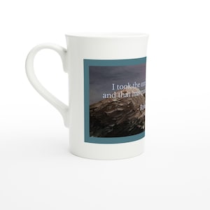 May include: White ceramic mug with a curved handle. The mug features a rectangular image of a mountain range with text that reads "I took the one and that has made all the difference." A quote by Robert Frost.