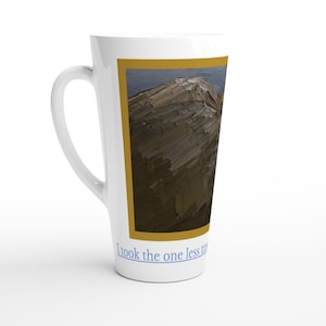 May include: White latte mug with a curved handle. The mug features a rectangular image of a mountain landscape in shades of brown and blue, framed by a gold border. The text "I took the one less" is printed at the bottom.
