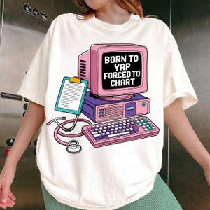 Puede incluir: Camiseta color crema con un gráfico de ordenador retro en rosa y morado, con el texto "BORN TO YAP FORCED TO CHART". También se muestra una carpeta y un estetoscopio. La camiseta es de estilo casual y unisex.