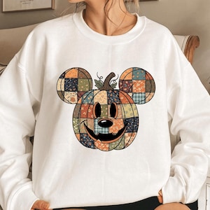 May include: White sweatshirt featuring a pumpkin design with Mickey Mouse ears. The pumpkin is made of patchwork fabric in orange, blue, and green. The design has a smiling face and a stem with leaves. A fun, festive top for fall.