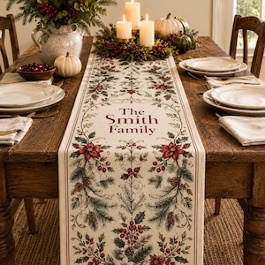 May include: A cream-colored fabric table runner featuring a festive botanical print of red poinsettias, green pine branches, pinecones, and holly berries. The center of the runner displays the personalized text The Smith Family in red.