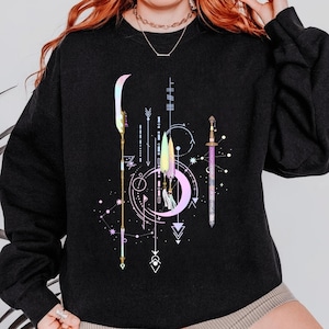 Puede incluir: Sudadera negra con un diseño celestial que presenta una luna creciente, flechas y una espada. El gráfico incluye formas geométricas y un patrón similar a una constelación en colores pastel. El diseño tiene una estética mística y bohemia.