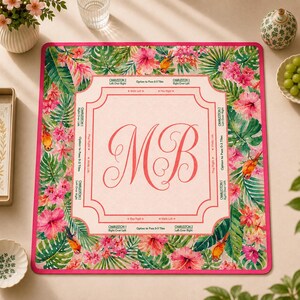 May include: A square mahjong mat featuring a tropical floral pattern with pink hibiscus flowers, green palm leaves, and orange birds. The center has a light pink square with the monogram MB and game instructions printed in black text.