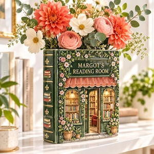 Könnte beinhalten: Eine dunkelgrüne, buchförmige Keramikvase mit einer detaillierten Ladenfassade, dem Schriftzug "MARGOT'S READING ROOM" über einer rosa Markise, verziert mit skulpturalen Blumenranken und gefüllt mit einem Strauß aus rosa und weißen Blumen.