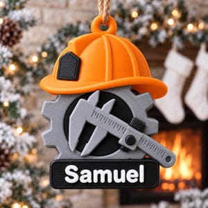 Op de afbeelding: Een hangend ornament met een oranje veiligheidshelm bovenop een grijs tandwiel en een schuifmaat. Onder het tandwiel bevindt zich een zwart rechthoekig plaatje met de naam Samuel in witte letters. Het ornament hangt aan een touwtje.