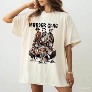 Puede incluir: Camiseta color crema con la inscripción "MURDER GANG" sobre un gráfico de seis personajes disfrazados. El gráfico presenta varios colores y detalles, con algunos personajes arrodillados. La camiseta es de estilo casual y oversize.