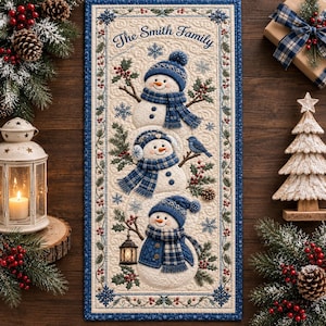 Könnte beinhalten: Ein rechteckiger, gesteppter Wandbehang mit drei übereinander gestapelten Schneemännern in blau karierten Mützen und Schals sowie dem Schriftzug The Smith Family. Das Design zeigt Stechpalmen, Beeren, Schneeflocken und eine Laterne.