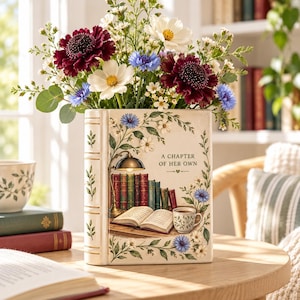 Könnte beinhalten: Eine cremefarbene Keramikvase in Buchform mit einer aufgedruckten Illustration eines Schreibtisches mit Büchern, einer Lampe und einer Tasse. Der Text A CHAPTER OF HER OWN ist auf der Vorderseite aufgedruckt. Die Vase ist mit dunkelroten und weißen Blumen gefüllt.