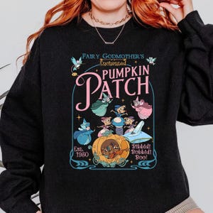 Pode incluir: Camisola preta com um gráfico colorido de "Fairy Godmother's Enchanted Pumpkin Patch". O design inclui fadas de desenhos animados, uma carruagem de abóbora e a frase "Bibbidi Bobbidi Boo!" com o ano "Est. 1980."