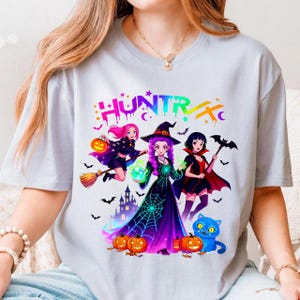 May include: A light grey t-shirt with a colourful graphic of three cartoon witches, pumpkins, bats, and a blue cat. The word "HUNTR/X" is printed above the witches in a rainbow gradient.