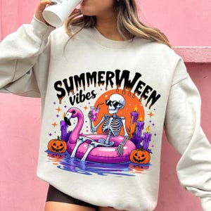 Op de afbeelding: Een lichtbeige sweatshirt met een kleurrijke afbeelding. De afbeelding toont een skelet met een zonnebril, ontspannen op een roze flamingo-drijver, met een drankje, met de tekst "Summerween vibes". Pompoenen, kaarsen en sterren maken het ontwerp compleet.