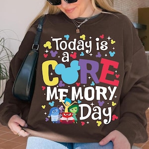 May include: A brown sweatshirt featuring the phrase "Today is a CORE MEMORY Day" in vibrant colours. The design incorporates Mickey Mouse ears and cartoon characters. The sweatshirt is a relaxed fit.