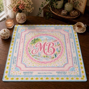 May include: A square Mahjong mat featuring a central pink monogram MB surrounded by a painted scene of buildings and palm trees. The border includes text instructions like Charleston 1, Charleston 2, Walls Left, and Option to Pass 0-3 Tiles.