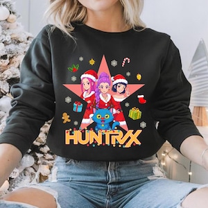 Puede incluir: Sudadera negra con un diseño navideño. Presenta tres mujeres de estilo anime con gorros de Papá Noel, un gato azul y la palabra "HUNTRAX" en dorado. El diseño está rodeado de copos de nieve y regalos festivos.