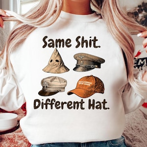 Op de afbeelding: Witte sweatshirt met de tekst "Same Shit. Different Hat." met illustraties van vier verschillende hoeden: een puntmuts, een militaire pet, een platte pet en een oranje baseballpet met tekst.