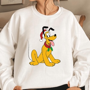 Puede incluir: Sudadera blanca con un diseño de dibujos animados de Pluto, el perro. El perro amarillo tiene orejas negras, lleva un gorro de Papá Noel y un collar rojo con acebo. El diseño está centrado en la parte delantera de la sudadera.