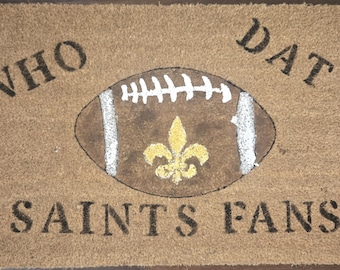 May include: A rectangular doormat with the words "WHO DAT SAINTS FANS" in black block letters. A brown football with white stripes and a gold fleur-de-lis is in the center.
