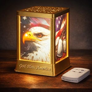 Puede incluir: Una linterna rectangular dorada con temática patriótica. La linterna presenta una imagen de un águila calva y la bandera estadounidense. Las palabras "God Bless America" están inscritas en la base. Un pequeño mando a distancia blanco está cerca.