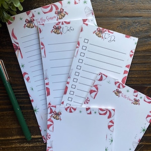 Puede incluir: Un conjunto de blocs de notas de papel blanco con temática navideña, decorados con rayas de bastones de caramelo rojos y blancos, campanas doradas y hombres de jengibre. Incluye hojas rayadas, una lista de tareas y notas cuadradas.