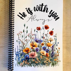 May include: A spiral-bound notebook with a white cover. The cover has the text "He is with you Always" in black script, above a watercolour illustration of colourful wildflowers in orange, purple, blue, and pink.