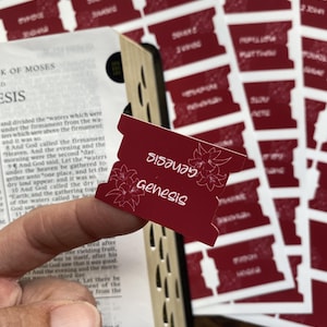 May include: A set of deep red adhesive bible index tabs featuring white floral line art and text. Each tab displays the name of a specific book of the Bible in a white script font. The tabs are presented on a sheet and one is shown applied to a page.