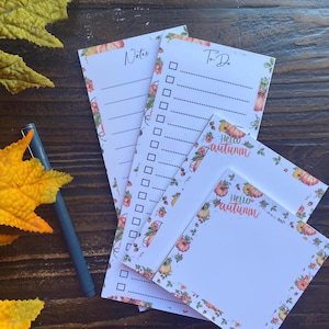 May include: A set of paper stationery including two long vertical notepads labeled Notes and To Do, and two square sticky notes with the text Hello Autumn. All items feature a decorative border of pumpkins and foliage. A gray pen is included.