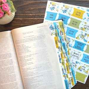 May include: An open book with the word "Genesis" visible, alongside colorful Bible tabs with floral and butterfly designs. A small pot of pink roses sits in the background, creating a soft, inviting aesthetic. The tabs are ready to be placed in the book.