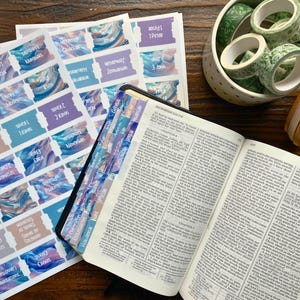 May include: An open Bible with colorful tab stickers and sheets of Bible verse labels. Decorative green washi tape and a wooden bookmark are also present. The labels and tabs have a watercolor design.