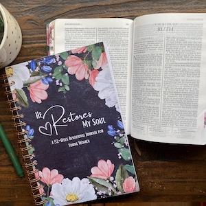 May include: A spiral-bound devotional journal with a dark background featuring pink and white floral illustrations and the text He Restores My Soul A 52-Week Devotional Journal For Young Women, positioned next to an open Bible with colorful page tabs.
