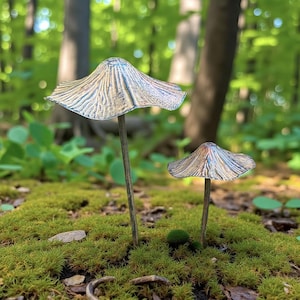 May include: Two decorative metal mushrooms with textured caps in shades of silver, blue, and gold stand in a bed of green moss. The stems are slender and brown. The background is a blurred forest scene with trees and foliage.
