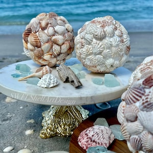 May include: Two decorative spheres covered in seashells, displayed on a marble-topped stand with a gold base. The spheres are in shades of brown and white. Sea glass and seashells are scattered around the stand, with the ocean in the background.