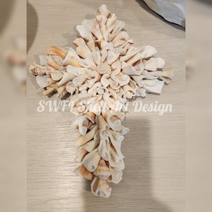 May include: A cross-shaped wall decoration constructed from various white and tan seashells layered to create a textured surface. The center features a small spiral shell, and the text SWFL Shell Art Design is overlaid across the middle.