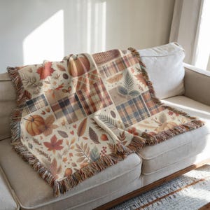 May include: A beige throw blanket with a patchwork design featuring pumpkins, leaves, and plaid patterns in shades of orange, brown, and cream. The blanket has a fringed edge and is draped over a beige sofa, creating a cozy autumn theme.
