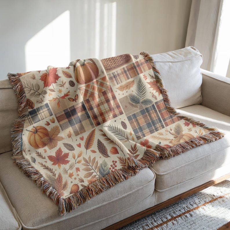 May include: A beige throw blanket with a patchwork design featuring pumpkins, leaves, and plaid patterns in shades of orange, brown, and cream. The blanket has a fringed edge and is draped over a beige sofa, creating a cozy autumn theme.